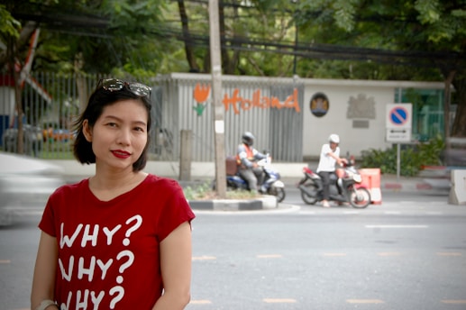 A woman wearing a red t-shirt with the text 'WHY? WHY? WHY?' is standing on a street. Behind her, two people on scooters are visible, parked near the road. There's a metal fence with the word 'Holland' written on it in orange letters and a sign indicating no parking. Trees in the background create a green, natural setting.
