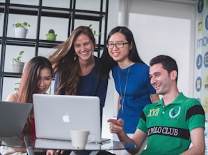 A group of four young adults are gathered around a laptop, smiling and engaging with the content on the screen. They appear to be in a modern office or meeting space with a shelf holding plants in the background. A coffee cup is visible on the table next to the laptop.