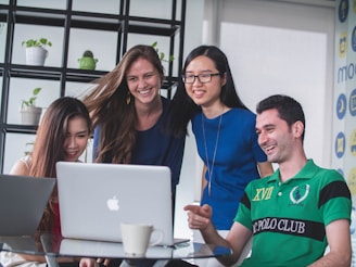 A group of four young adults are gathered around a laptop, smiling and engaging with the content on the screen. They appear to be in a modern office or meeting space with a shelf holding plants in the background. A coffee cup is visible on the table next to the laptop.