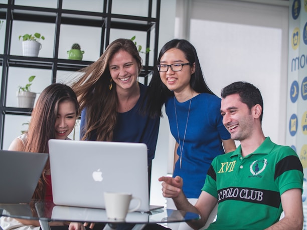 A group of four young adults are gathered around a laptop, smiling and engaging with the content on the screen. They appear to be in a modern office or meeting space with a shelf holding plants in the background. A coffee cup is visible on the table next to the laptop.