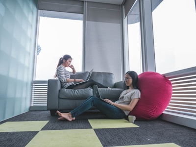 Two people are working on laptops in a modern room with large windows. One person is sitting on a sofa, while the other is sitting on the floor propped against a large pink beanbag. The room features a minimalist design with a checkered carpet and muted colors.