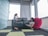Two people are working on laptops in a modern room with large windows. One person is sitting on a sofa, while the other is sitting on the floor propped against a large pink beanbag. The room features a minimalist design with a checkered carpet and muted colors.