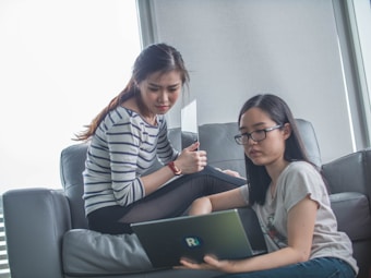 Two women are sitting on a sofa. One woman is using a laptop, while the other is holding a tablet and looking on. The setting appears relaxed and casual.