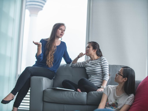 Three people are sitting on a grey couch in a modern, bright room. One individual, wearing a blue blouse, is gesturing with their hand while speaking to two others. The two listeners are sitting casually on the couch; one is wearing a striped shirt and appears to be pondering, while the other, in a graphic tee and glasses, looks on attentively. In the background, a blurred structure is visible through the window, suggesting an urban setting.