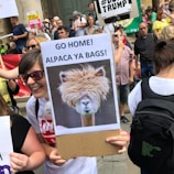 A crowd of people at a protest holding various signs. One person in the foreground is smiling and holding a humorous sign that features an alpaca with the text 'Go Home! Alpaca Ya Bags.' Other protesters and signs are visible in the background, contributing to the lively atmosphere.