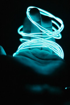 A hand is outstretched in a dark environment, wrapped with a glowing neon blue wire that emits a soft, luminous light.