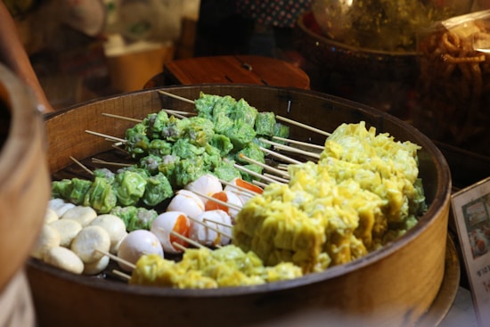 A wooden steamer basket filled with skewers of colorful dumplings and eggs. The dumplings are predominantly green and yellow, and there are pieces of boiled eggs arranged together.