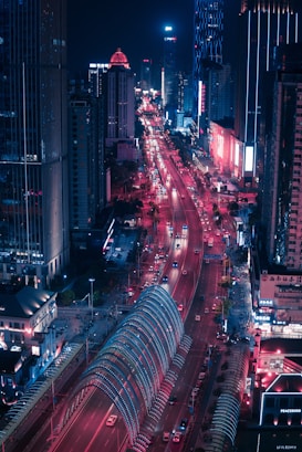 An urban nightscape featuring a busy city street illuminated with vibrant red and blue neon lights. High-rise buildings line the street, which is bustling with vehicles. A distinctive arched structure with blue and red lights spans across the road, creating a visually striking effect.