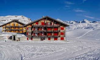Two alpine chalet-style buildings with colorful shutters are situated among snowy landscapes and mountains under a clear blue sky. Snow tracks lead toward the buildings, and the surrounding area appears pristine and undisturbed.