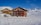 Two alpine chalet-style buildings with colorful shutters are situated among snowy landscapes and mountains under a clear blue sky. Snow tracks lead toward the buildings, and the surrounding area appears pristine and undisturbed.
