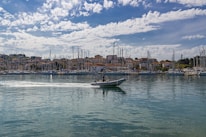 A peaceful harbor scene with numerous sailboats anchored in a marina. In the foreground, a small motorboat with a person aboard creates a gentle wake as it moves across the calm water. A backdrop of charming hillside buildings sits under a partly cloudy sky, adding depth to the coastal setting.