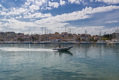 A peaceful harbor scene with numerous sailboats anchored in a marina. In the foreground, a small motorboat with a person aboard creates a gentle wake as it moves across the calm water. A backdrop of charming hillside buildings sits under a partly cloudy sky, adding depth to the coastal setting.