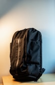 A black backpack with a sleek design is resting upright on a light wooden surface. The background features a soft gradient from light gray to off-white, highlighting the bag's texture and contour. The backpack has a prominent front zipper and multiple compartments, showcasing its functionality and minimalist style.