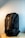 A black backpack with a sleek design is resting upright on a light wooden surface. The background features a soft gradient from light gray to off-white, highlighting the bag's texture and contour. The backpack has a prominent front zipper and multiple compartments, showcasing its functionality and minimalist style.