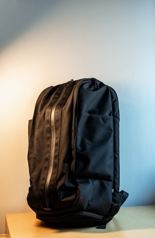 A black backpack with a sleek design is resting upright on a light wooden surface. The background features a soft gradient from light gray to off-white, highlighting the bag's texture and contour. The backpack has a prominent front zipper and multiple compartments, showcasing its functionality and minimalist style.