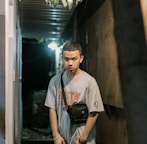 A young person with short hair stands in a narrow corridor at night, wearing a gray t-shirt with lettering and a black crossbody bag. The corridor is dimly lit by an overhead light, casting shadows along the walls. There are potted plants visible hanging from the ceiling.