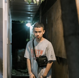 A young person with short hair stands in a narrow corridor at night, wearing a gray t-shirt with lettering and a black crossbody bag. The corridor is dimly lit by an overhead light, casting shadows along the walls. There are potted plants visible hanging from the ceiling.