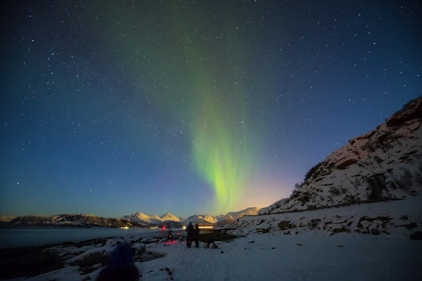 Two people on a snowy shore watching a green aurora band above the fjord mountains near Tromsø