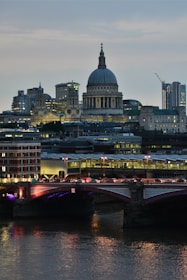 A prominent cityscape featuring an iconic cathedral with a large dome, surrounded by modern skyscrapers and historic architecture. The foreground shows a bridge spanning a river, illuminated by streetlights and reflecting in the water below.