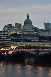 A prominent cityscape featuring an iconic cathedral with a large dome, surrounded by modern skyscrapers and historic architecture. The foreground shows a bridge spanning a river, illuminated by streetlights and reflecting in the water below.