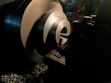 A metal lathe is working on shaping a cylindrical piece, with sharp edges visible and metal shavings scattered around. The lathe is in a dimly lit workshop environment, with the machinery casting dark shadows.