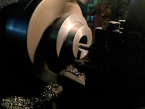 A metal lathe is working on shaping a cylindrical piece, with sharp edges visible and metal shavings scattered around. The lathe is in a dimly lit workshop environment, with the machinery casting dark shadows.