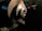 A metal lathe is working on shaping a cylindrical piece, with sharp edges visible and metal shavings scattered around. The lathe is in a dimly lit workshop environment, with the machinery casting dark shadows.