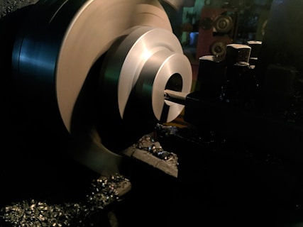 A metal lathe is working on shaping a cylindrical piece, with sharp edges visible and metal shavings scattered around. The lathe is in a dimly lit workshop environment, with the machinery casting dark shadows.