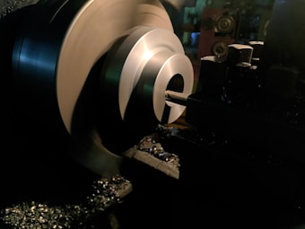 A metal lathe is working on shaping a cylindrical piece, with sharp edges visible and metal shavings scattered around. The lathe is in a dimly lit workshop environment, with the machinery casting dark shadows.