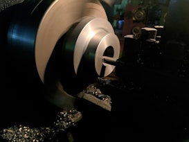 A metal lathe is working on shaping a cylindrical piece, with sharp edges visible and metal shavings scattered around. The lathe is in a dimly lit workshop environment, with the machinery casting dark shadows.