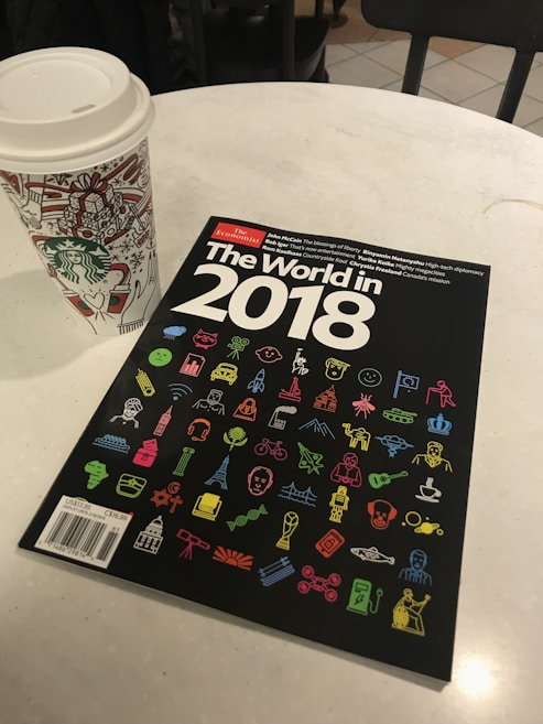 A magazine titled 'The World in 2018' from The Economist is placed on a round white table. The cover is black and features numerous colorful icons related to different themes like technology, travel, and entertainment. Next to the magazine is a disposable coffee cup with decorative holiday illustrations, likely from Starbucks.