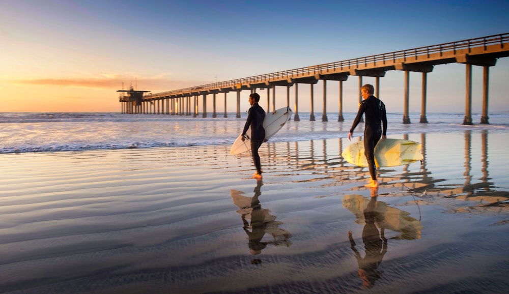 two person walking on the seaside holding surfboards