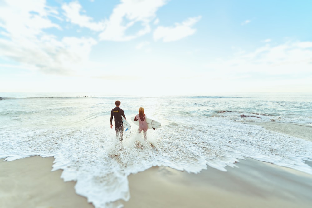 candid photography of man and woman at seashore carrying surfboards under cirrus clouds and blue calm sky
