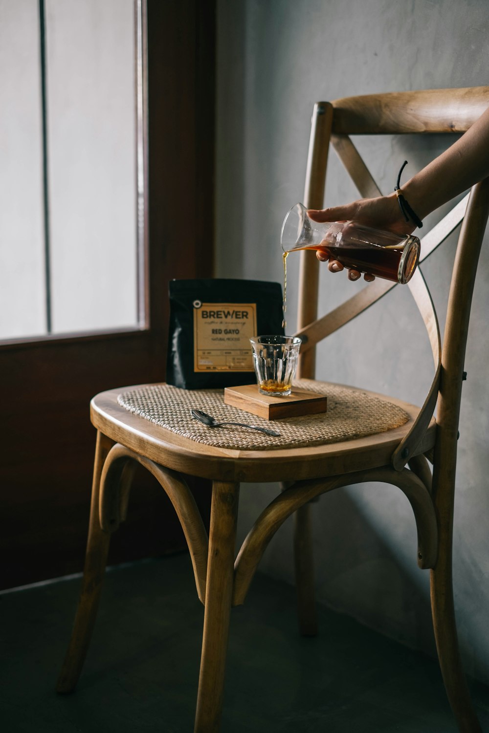 Person mit klarer Glaskaraffe, die einen Whisky in einen klaren Trinkglasbecher auf einem braunen Holzstuhl in der Nähe des Klarglasfensters im Zimmer gießt