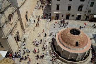 A bustling historic square is filled with tourists walking and exploring. The square is lined with old stone buildings featuring arched windows and detailed facades. A large, circular stone fountain with an intricate design stands prominently in the foreground. The ground is paved with light-colored stones, creating a uniform appearance. People of various ages are scattered throughout, some holding umbrellas to shield themselves from the sun.