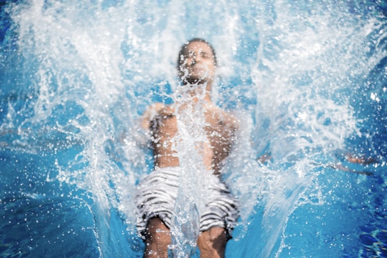 A person is plunging into water, creating a large splash. The water is clear and blue, and the individual appears to be wearing striped shorts. The image is dynamic and conveys motion.