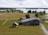 A scenic rural landscape featuring traditional wooden buildings, green fields, and a lake in the background. The architecture of the buildings reflects a rustic style. Trees are scattered throughout the area, adding to the serene and natural atmosphere.