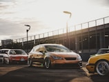 Several modified cars are parked on a paved area with a modern, urban backdrop featuring a metal fence and streetlights. The sun sets in the background, casting warm light and long shadows across the scene.