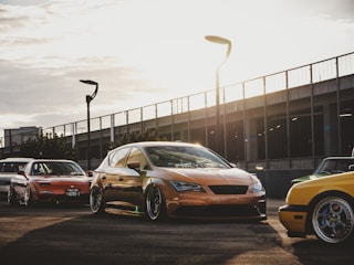 Several modified cars are parked on a paved area with a modern, urban backdrop featuring a metal fence and streetlights. The sun sets in the background, casting warm light and long shadows across the scene.