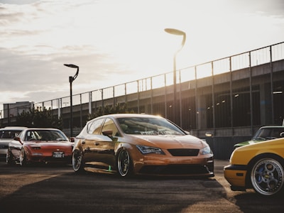 Several modified cars are parked on a paved area with a modern, urban backdrop featuring a metal fence and streetlights. The sun sets in the background, casting warm light and long shadows across the scene.