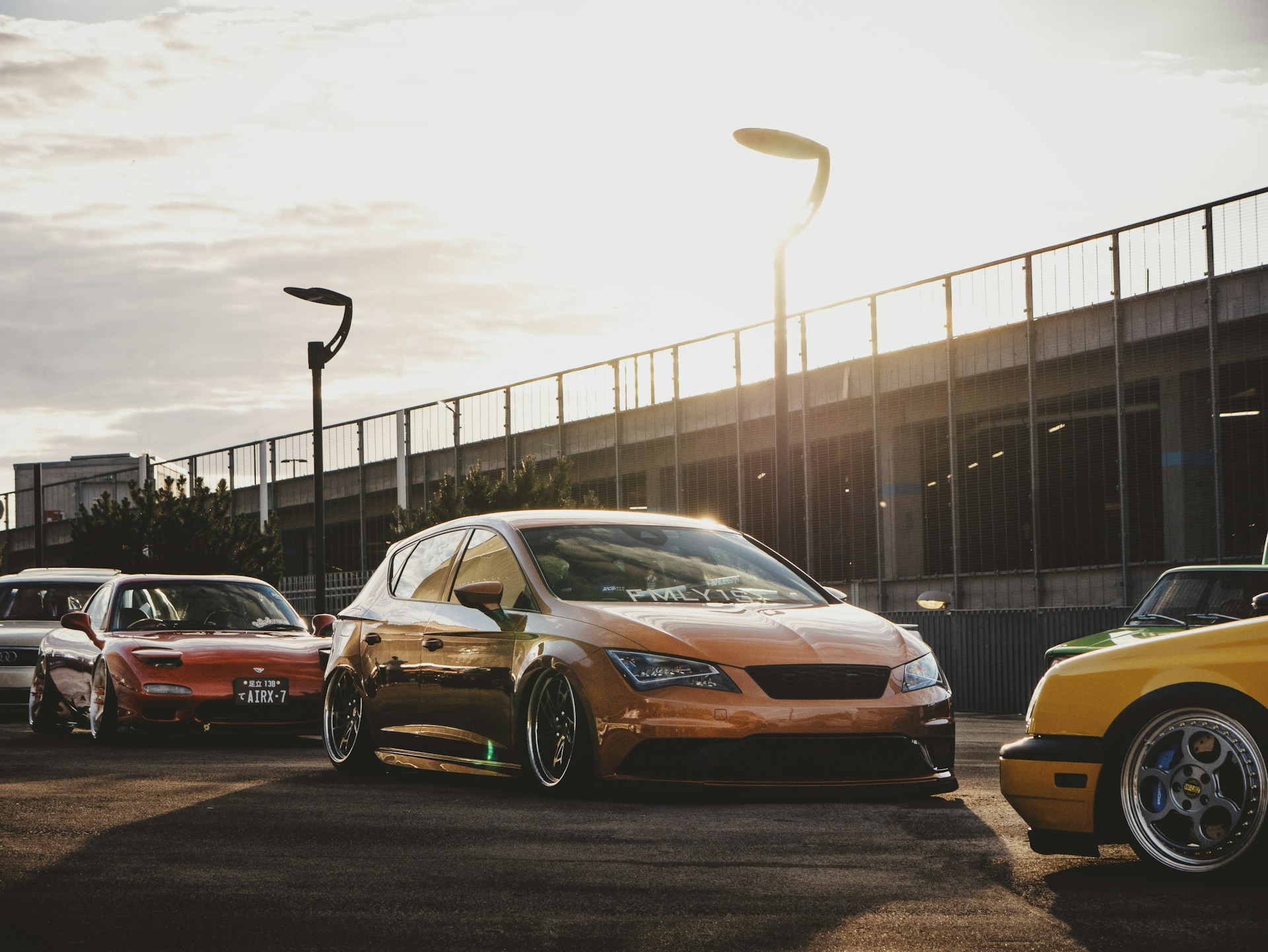Several modified cars are parked on a paved area with a modern, urban backdrop featuring a metal fence and streetlights. The sun sets in the background, casting warm light and long shadows across the scene.