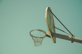 A vintage-style basketball hoop with a rusted metal frame and a worn net against a clear blue sky. The backboard is simple and slightly weathered, suggesting years of use.