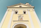 The image features the facade of a yellow church building with a prominent arched entrance and decorative white trim. Above the entrance, there is a colorful religious mural depicting saints or biblical figures. The architecture includes arched windows and detailed cornices. The steeple has a cross at the top and is partially visible against a clear blue sky.