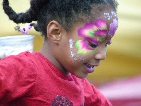 A young girl with colorful face paint, featuring vibrant pink, green, and purple flower designs on her face. She is wearing a red sweater and has her hair styled in braids with a pink hair tie.
