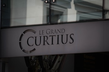 The image features a building facade with the name 'Le Grand Curtius' prominently displayed. The text is set in an elegant serif font, and above it is a circular emblem. The background consists of glass elements and architectural details, suggesting an institutional or cultural establishment.