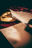 A hand is writing on a piece of paper with text that reads 'be happy for this moment, this moment is Life.' The paper is placed on a red surface with a decorative pattern. A couple of bananas and other papers are beside the main sheet. The lighting creates a warm and intimate atmosphere.