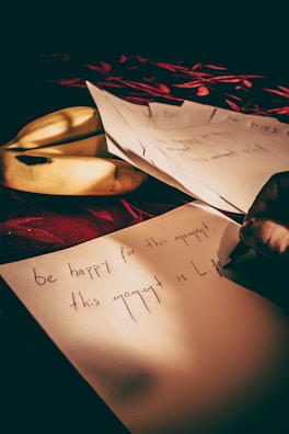 A hand is writing on a piece of paper with text that reads 'be happy for this moment, this moment is Life.' The paper is placed on a red surface with a decorative pattern. A couple of bananas and other papers are beside the main sheet. The lighting creates a warm and intimate atmosphere.