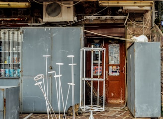 A weathered storefront with a wooden door displaying posters and advertisements. An air conditioning unit is mounted above. Various metal frames and stands are placed in front of the door. A white cat lounges on top of a metal cabinet to the right, while a tabby cat sits on the ground near the door.