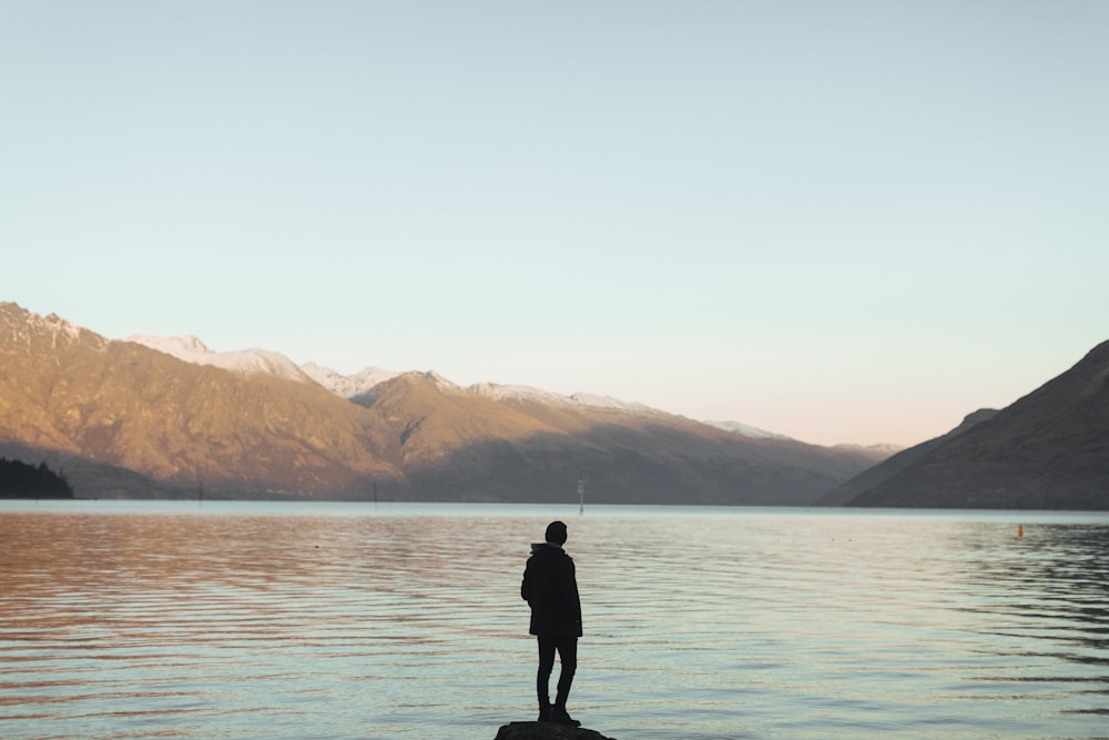 silhouette of person near calmly body of water over mountain under blue sky during daytime