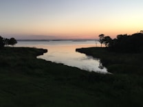 A tranquil lakeside scene at dusk with calm water reflecting the soft colors of the evening sky. Silhouetted trees frame the edges of the water, adding a sense of serenity and natural beauty.